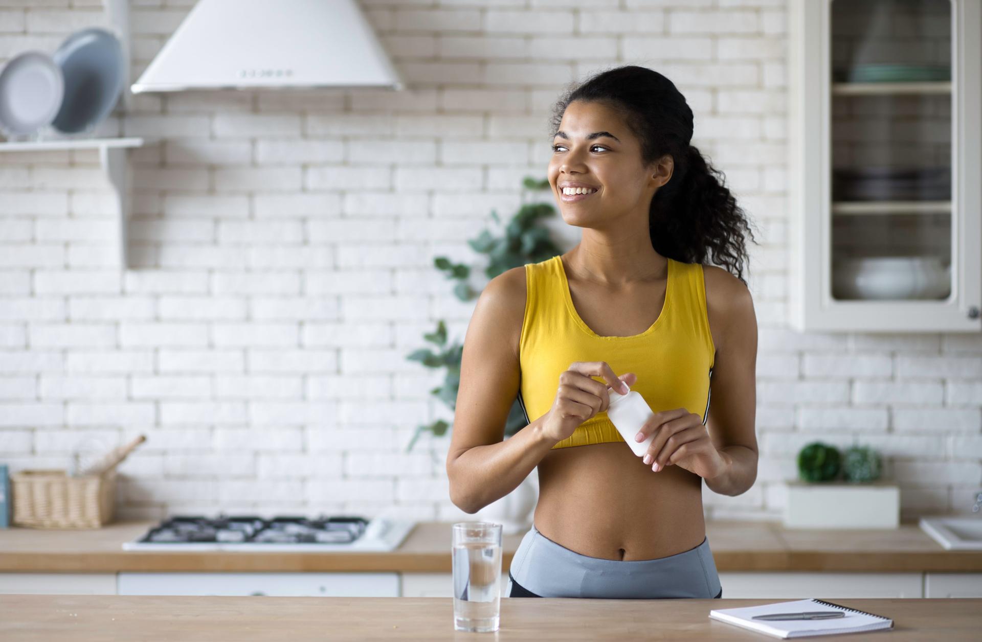 Happy sportive african american woman in sportswear holding a bottle of nutritional supplements, looking happily out the window, healthy lifestyle